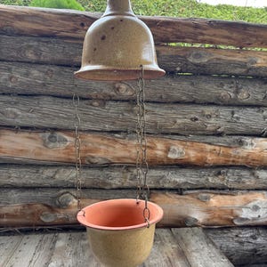 Op de afbeelding: Een hangende plantenbak met een klokvormige bovenkant en een ronde pot, verbonden door kettingen. De plantenbak heeft een gespikkelde bruine kleur met een oranje binnenkant. De achtergrond toont een houten hek en een houten oppervlak.