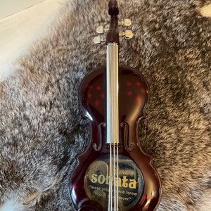 May include: A dark red toy violin with a silver string and a gold label that reads "sonata please play in piece forme you good player".