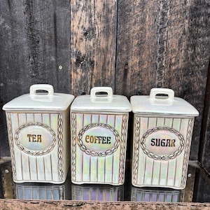 May include: Three vintage ceramic tea, coffee, and sugar canisters. Each has a lid and handle. The canisters are white with vertical stripes and gold trim. The words "TEA", "COFFEE", and "SUGAR" are displayed in gold. A classic kitchen set.