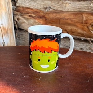 May include: A white ceramic mug featuring a graphic illustration of a green-skinned character with bright orange hair, a stitched forehead scar, and a friendly facial expression set against a black background with white speckles.