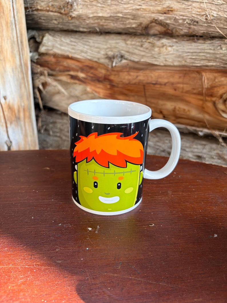 May include: A white ceramic mug featuring a graphic illustration of a green-skinned character with bright orange hair, a stitched forehead scar, and a friendly facial expression set against a black background with white speckles.
