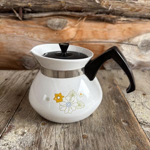 May include: White ceramic teapot with a black lid and handle. The teapot features a floral design in yellow, blue, and green. A silver band encircles the middle of the teapot. The teapot is set on a wooden surface.