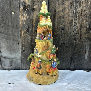 May include: A multi-level Christmas nativity scene with a brown, sandy base. The scene features a white building with a gold dome, a stable with figures of Mary, Joseph, and baby Jesus, and three wise men riding camels.