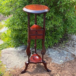 May include: A dark brown wooden pedestal stand featuring a circular top, three barley twist legs, and two small stacked drawers positioned in the center. The base includes a triangular shelf with a central circular indentation and curved feet.