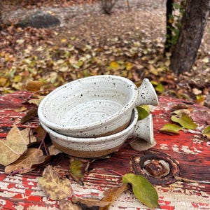 Può includere: Due ciotole in ceramica fatte a mano con interno bianco maculato e base marrone. Ogni ciotola ha un piccolo manico. Le ciotole sono impilate l'una sull'altra. Le ciotole sono appoggiate su una superficie rossa con vernice scrostata, circondate da foglie cadute.