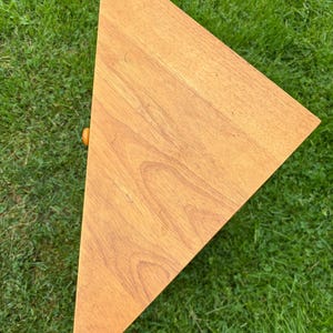 May include: A triangular wooden shelf with a light brown wood grain finish. The shelf features a flat top surface and a small, round, orange-coloured knob protruding from the side edge, indicating a drawer or compartment front.