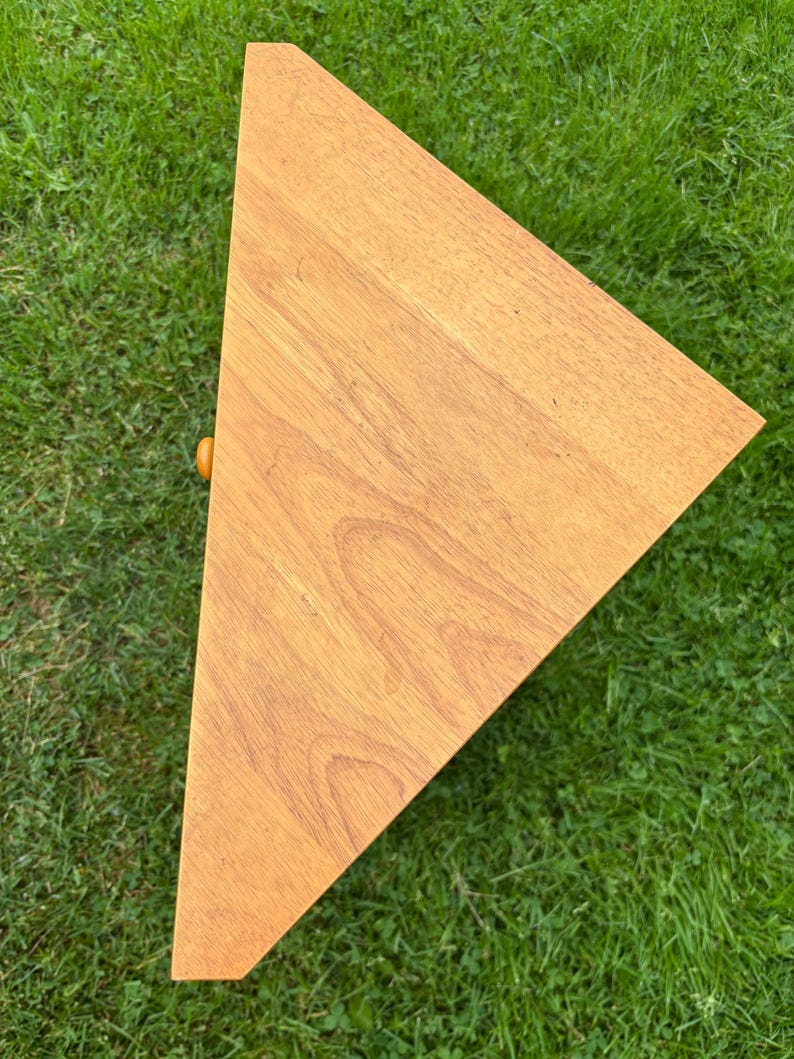 May include: A triangular wooden shelf with a light brown wood grain finish. The shelf features a flat top surface and a small, round, orange-coloured knob protruding from the side edge, indicating a drawer or compartment front.