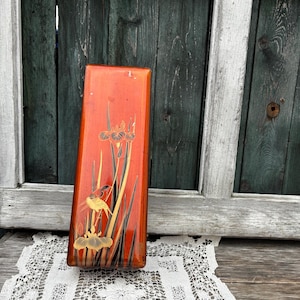 Può includere: Una scatola rettangolare in legno con finitura laccata rossa e dettagli dorati. La scatola presenta un design di ispirazione giapponese con un uccello e fiori di iris.