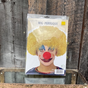 May include: A yellow curly wig with a red clown nose and a white face with blue and black makeup. The packaging says "Wig - Perruque".