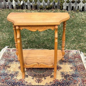 May include: A light brown, antique-style side table with a curved top and a lower shelf. The table has decorative carved details and turned legs. It is set on a patterned rug, with a white picket fence in the background.