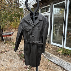 以下が含まれることがあります： ボタン留めとベルトが付いた茶色の革のトレンチコート。コートは黒いスタンドにかけられています。