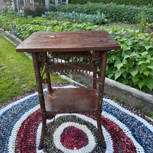 May include: A brown, vintage side table with a rectangular top and a lower shelf. The table has detailed woven elements and stands on four legs. It is placed on a colourful, circular rug with red, white, and blue stripes. A garden is visible in the background.