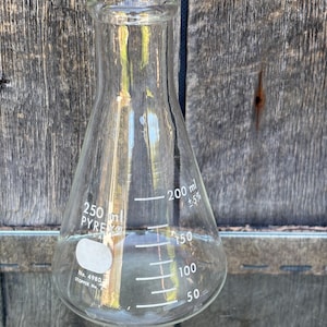 Puede incluir: Un matraz Erlenmeyer Pyrex transparente de 250 ml con marcas de medición blancas. El matraz tiene una base ancha y un cuello estrecho, con una etiqueta que dice "No. 4980 Stopper No.". El matraz está contra un fondo de madera desgastada.