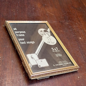 May include: A rectangular gold-toned metal picture frame with an ornate embossed border. The glass insert features black and white graphics of a couple and a baby, with text reading all purpose frame, 5x7, and Columbia Frame Inc.