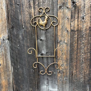 Può includere: Decorazione murale in metallo color oro con un design a gallo. Il pezzo decorativo ha un design ornato a forma di voluta con una forma rettangolare ed è montato su uno sfondo di legno invecchiato.