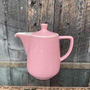 Puede incluir: Una tetera de cerámica rosa con tapa blanca y asa. La tetera está sobre una superficie de madera.