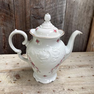 Puede incluir: Tetera de porcelana blanca con diseño floral. La tetera presenta un asa curva, un pico vertedor y una tapa con un adorno decorativo. El cuerpo está adornado con volutas en relieve y pequeños detalles florales rosas.
