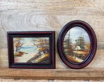 Op de afbeelding: Twee ingelijste schilderijen met landschappen. Het schilderij aan de linkerkant is in een rechthoekige lijst en toont een meer en bomen in herfstkleuren. Het schilderij aan de rechterkant is in een ovale lijst en toont een hut in het bos.