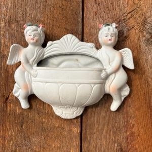 Könnte beinhalten: Ein weißer Keramik-Wandtopf mit zwei Cherubfiguren auf jeder Seite. Der Topf hat einen gewellten Rand und ein Blumenmuster.