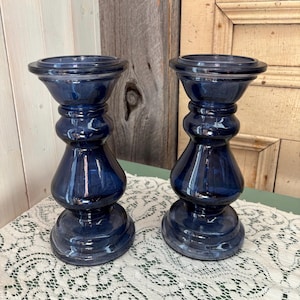 May include: Two navy blue glass candle holders with a bulbous design. The candle holders have a wide base, a central section with rounded details, and a flared top. They are placed on a white lace tablecloth.