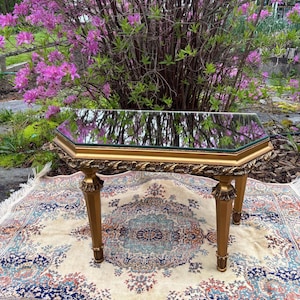 May include: An antique-style, gold-coloured side table with a mirrored top. The table has an octagonal shape and decorative carvings. It rests on four legs and is set on a patterned rug, with flowering bushes in the background.