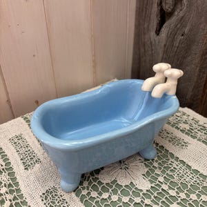 Puede incluir: Una bañera de cerámica azul claro con detalles de grifos blancos. La bañera en miniatura tiene cuatro patas pequeñas y se exhibe sobre una superficie ribeteada con encaje. El fondo presenta tablones de madera.