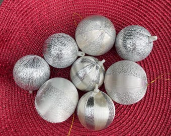Puede incluir: Una colección de adornos navideños plateados y blancos con diversas texturas y diseños. Los adornos están dispuestos sobre una superficie roja texturizada, creando una exhibición festiva. Cada adorno tiene una cuerda dorada para colgar.