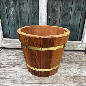 Könnte beinhalten: Ein Holzeimer mit konischem Design, der aus vertikalen Holzstäben besteht. Der Eimer ist von zwei Messingbändern umschlossen. Das Holz hat einen warmen, natürlichen Farbton, und die Messingbänder sorgen für einen Hauch von Kontrast. Der Eimer dient wahrscheinlich dekorativen oder Aufbewahrungszwecken.