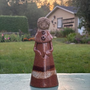 May include: A ceramic figurine of a person with a brown dress and a light brown head. The figurine has a round head with short hair and a small face. The dress has a layered design with a white stripe. Two brown buttons are on the chest.