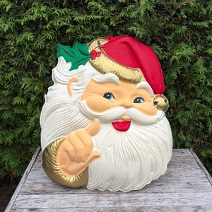May include: A moulded plastic Santa Claus wall decoration featuring a red hat with a gold band, a white beard, and a green holly sprig with red berries. The figure has a peach-coloured face, blue eyes, red lips, and a gold-coloured glove giving a thumbs up.