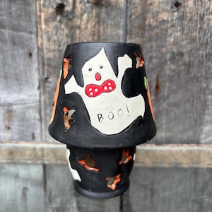 Peut inclure: Un bougeoir d'Halloween en céramique noire avec un motif de fantôme blanc et le mot "BOO!". Le fantôme porte un nœud papillon rouge. Des découpes orange et noires de citrouilles et de chauves-souris décorent les côtés. Le bougeoir est sur une surface en verre.
