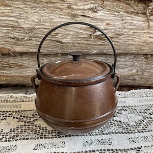 May include: A brown ceramic pot with a lid and a black metal handle. The pot has a rustic, vintage look.