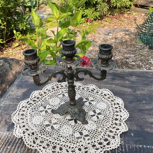 Peut inclure: Un chandelier en bronze antique à trois bougeoirs, posé sur un napperon en dentelle blanche. Le chandelier présente des détails ornés et est placé sur une surface en bois vieilli, avec de la verdure en arrière-plan.
