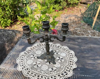 Puede incluir: Un candelabro antiguo de bronce con tres candeleros, colocado sobre un tapete de encaje blanco. El candelabro tiene detalles ornamentados y se coloca sobre una superficie de madera envejecida, con vegetación de fondo.
