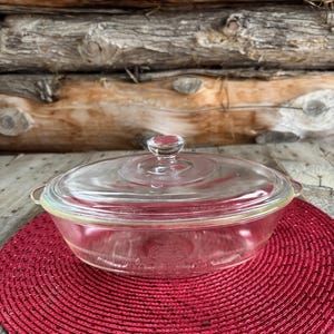 Puede incluir: Una fuente para horno de vidrio transparente con tapa a juego. El plato tiene forma redonda con dos asas y se asienta sobre un salvamanteles rojo tejido. El fondo presenta una pared de madera rústica.