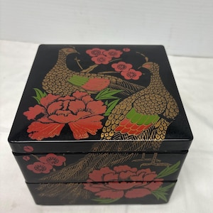Op de afbeelding: Een zwarte gelakte houten doos met een ontwerp met twee verdiepingen. De doos is versierd met een kleurrijk bloemen- en vogelmotief. De vogels zijn goud, groen en rood, en de bloemen zijn rood en roze.