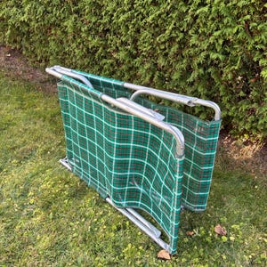 May include: A folded green and red plaid folding chair with a silver metal frame. The chair is sitting on green grass in front of a green hedge.