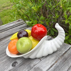 May include: A white ceramic cornucopia overflowing with colorful, glossy ceramic fruit. The arrangement includes a red apple, orange, green apple, purple plum, and yellow banana. The cornucopia is set on a weathered wooden surface.