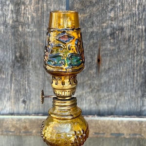 May include: A small amber glass oil lamp with a decorative floral design. The lamp has a brass wick holder and a clear glass chimney.