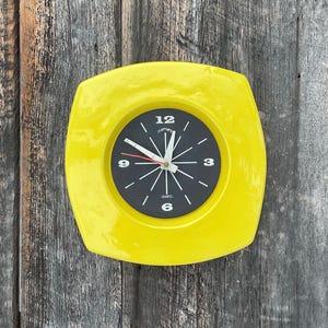 Op de afbeelding: Een gele vierkante klok met zwarte wijzers en een zwarte wijzerplaat. Op de wijzerplaat van de klok staan de woorden "moments" en "quartz" gedrukt.