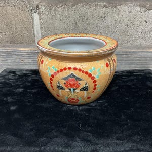 Puede incluir: Un pequeño macetero redondo de cerámica con un diseño floral marrón, amarillo, rojo y verde. El macetero tiene un interior blanco.