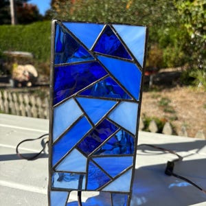 May include: A stained glass lamp with a geometric design in shades of blue. The lamp has a rectangular base and a cord.