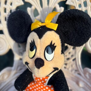 K&ouml;nnte beinhalten: Ein Vintage-Minnie-Mouse-Pl&uuml;schtier. Das Pl&uuml;schtier hat schwarze Ohren, eine gelbe Schleife und ein cremefarbenes Gesicht. Sie tr&auml;gt ein orangefarbenes Kleid mit wei&szlig;en Punkten. Das Spielzeug sitzt auf einem wei&szlig;en, verzierten Stuhl.
