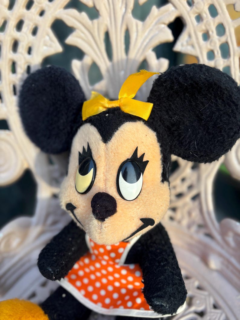 K&ouml;nnte beinhalten: Ein Vintage-Minnie-Mouse-Pl&uuml;schtier. Das Pl&uuml;schtier hat schwarze Ohren, eine gelbe Schleife und ein cremefarbenes Gesicht. Sie tr&auml;gt ein orangefarbenes Kleid mit wei&szlig;en Punkten. Das Spielzeug sitzt auf einem wei&szlig;en, verzierten Stuhl.