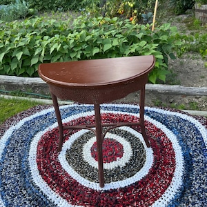 Peut inclure: Une table marron en demi-lune avec des détails tissés et trois pieds repose sur un tapis de chiffon circulaire coloré. Le tapis présente des cercles concentriques rouges, blancs, bleus et gris. Le fond montre un jardin avec des plantes vertes.