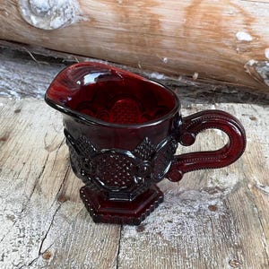 Peut inclure: Un pot à crème en verre rouge avec un motif texturé et une poignée. Le pot à crème a un pied.