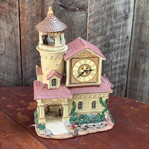 May include: A decorative clock shaped like a stone building with a tall tower, a red tiled roof, and a central clock face featuring Roman numerals and a compass star design. The structure includes arched windows, green vines, and small figures.