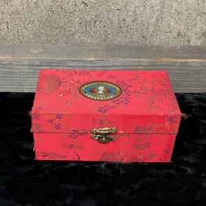 Può includere: Una scatola rossa con una chiusura dorata e un design a motivi blu e oro. La scatola ha un'etichetta con caratteri cinesi e il testo "HANDICRAFT FACTORY" in inglese.