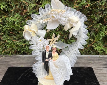 Op de afbeelding: Een vintage bruidstaarttopper met een bruid- en bruidegomfiguur. De bruid draagt een gele jurk en de bruidegom een zwart smoking. De topper is versierd met wit kant, bloemen en linten, en heeft een gouden filigraan basis.