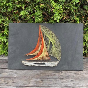 May include: A rectangular black board featuring a string art sailboat design. The boat hull is created with white string, while the sails are formed using orange and light yellow string wrapped around small metal nails to create geometric patterns.